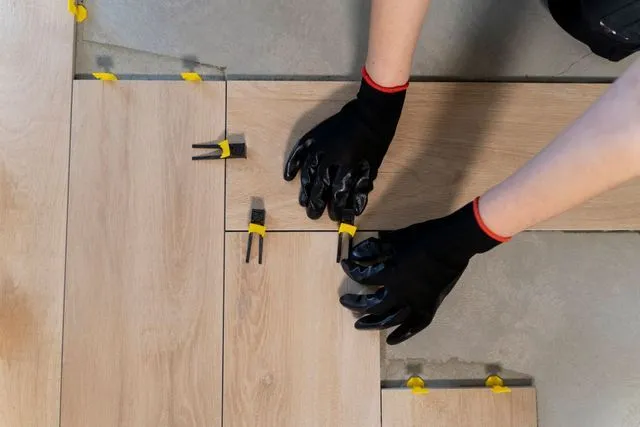 Hands in black protective gloves install wood-look floor tiles using yellow leveling spacers on a concrete surface.
