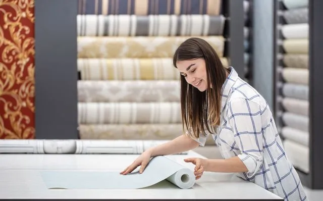 A person in a plaid shirt unrolls light-colored wallpaper on a white table in a store with shelves of wallpaper rolls.